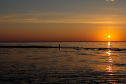 A lone fisherman casts a net from a sandbar at sunset in Batumi, Georgia, while several fishing rods stand planted in the shallow water as the sun reflects across the sea.