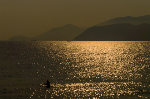 A lone swimmer stands in shimmering water as the sun dims during a partial solar eclipse, casting golden light across the sea and distant mountain silhouettes.