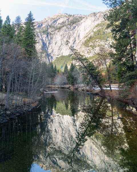 Granite cliffs and evergreen trees reflected in the calm waters of the Merced River in Yosemite Valley during winter.