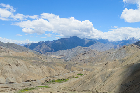 A wide view of Ladakh’s high-altitude desert shows rolling, barren mountains under a bright blue sky, with a narrow green river valley cutting through the landscape.