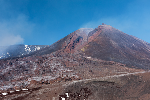 The collapsed summit cone of Mount Etna reveals fresh lava scars and ash-covered slopes, with patches of snow remaining on older flows.