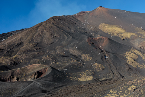 Dark volcanic slopes of Mount Etna rise against a clear blue sky, with ash, lava rock, and sparse vegetation marking past eruptions.