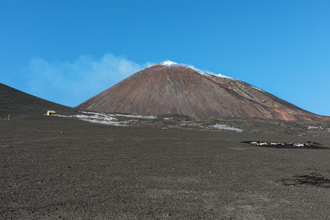 Mount Etna rises with its cone intact above a vast black lava field in Sicily, Italy, under a clear blue sky in February 2022.