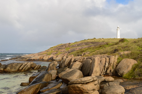 The Cape Leeuwin Lighthouse stands atop a rocky headland, with weathered granite boulders and waves breaking along the coastline under a cloudy sky.