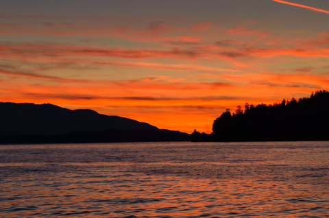 A vivid orange sunset glows above calm water in Southeast Alaska, with dark mountain and forested shoreline silhouettes against the sky.