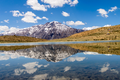 A calm alpine lake reflects snow-capped mountains and scattered clouds near Dhankar in the Spiti Valley, India.