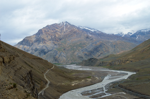 A braided river winds through the wide, barren Mudh Valley, surrounded by steep mountain slopes and snow-dusted peaks under an overcast sky.