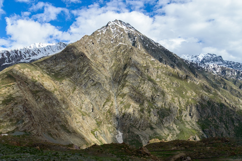 A steep, jagged mountain rises sharply in Ladakh, its rocky slopes streaked with snow near the summit beneath scattered clouds.