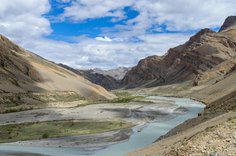 A pale blue glacial river winds through a broad valley in Ladakh, bordered by steep, barren mountains and patches of green vegetation under a partly cloudy sky.