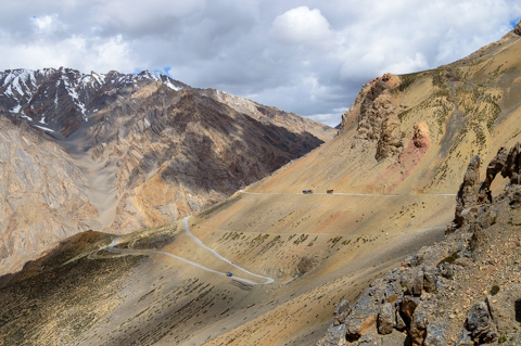 A winding mountain road cuts across the stark, rocky landscape of Ladakh, India, with barren slopes and distant snow-dusted peaks under a cloudy sky.
