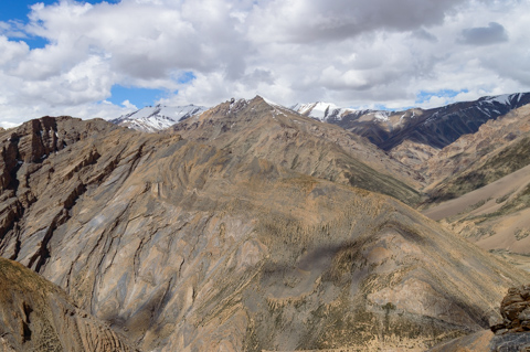Layered mountain ridges in Ladakh stretch across a high-altitude desert landscape, with pale rock faces and distant Himalayan peaks under a partly cloudy sky.