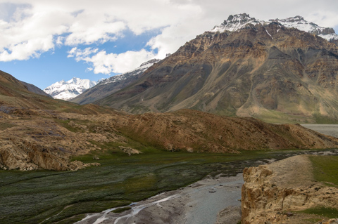 A glacial river flows through a rocky valley near Kaza in Himachal Pradesh, framed by barren slopes and snow-capped Himalayan peaks under a cloudy sky.