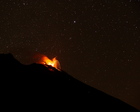 A wide-angle view shows Stromboli Volcano erupting at night, with glowing lava arcing into the air beneath a clear, star-filled sky.