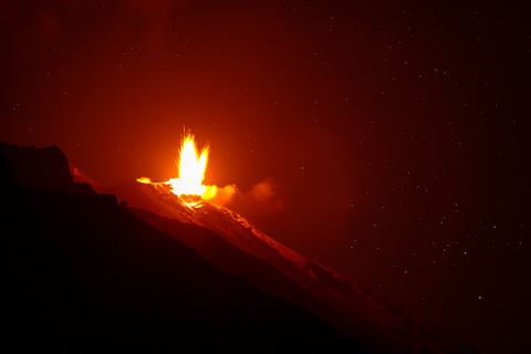 A bright lava fountain erupts from Stromboli Volcano at night, illuminating drifting smoke and the steep volcanic slope beneath a star-filled sky.
            