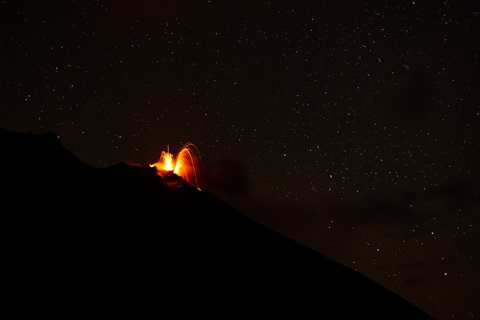 Lava arcs into the night sky as Stromboli Volcano erupts, glowing against a vast field of stars above the dark volcanic slope.