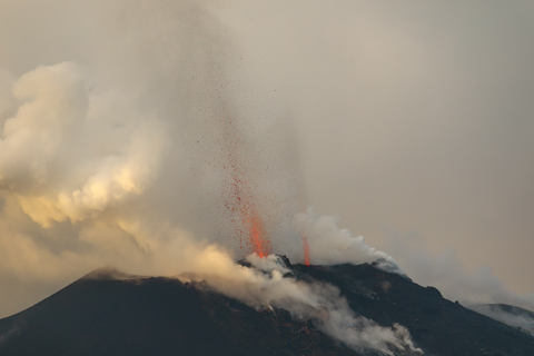 A close-up view of Stromboli Volcano erupting during the day, with ash and glowing lava jets rising through low clouds above the dark volcanic cone.