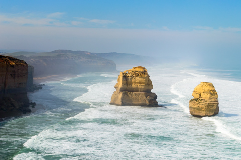 Two limestone sea stacks rise from the ocean as waves crash around them along a rugged coastline under clear skies.