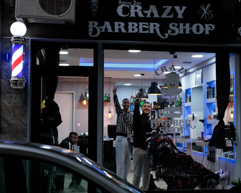 Two barbers pause and look toward the camera inside a neighborhood barbershop in Athens, Greece, as a customer sits in a barber chair.
            
