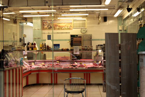An empty butcher shop in Athens, Greece, displays cuts of meat behind a glass counter under bright fluorescent lighting.
