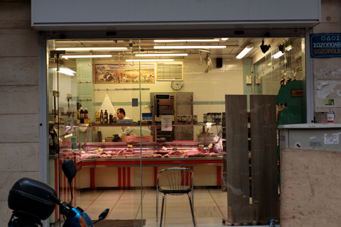 A butcher works behind a glass counter displaying cuts of meat inside a small neighborhood shop in Athens, Greece, seen from the street through the open storefront.
            