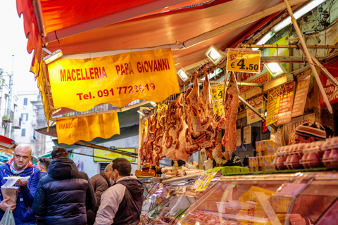 Customers stand at a meat stall at a street market in Palermo, Sicily, Italy, where cuts of meat hang beneath signs for Macelleria Papa Giovanni.