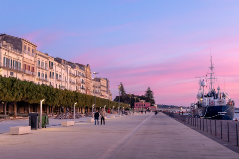 People walk along the seafront in Siracusa at sunset as the rescue vessel ResQ People is moored at the dock beneath a pink and purple sky.
