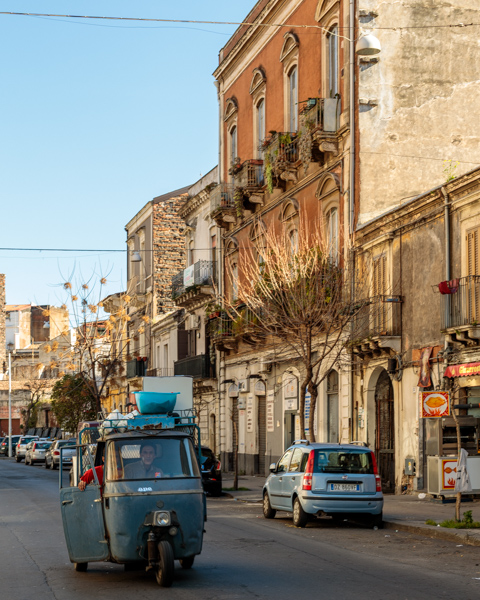 A three-wheeled tuk-tuk drives along a city street in Catania, Sicily, Italy, passing weathered buildings and parked cars in the historic center.