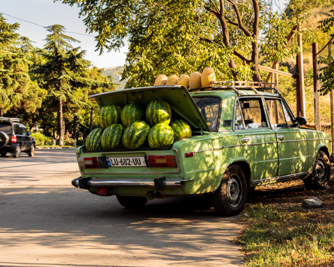 A vintage green car parked along a street in Tbilisi, Georgia, is filled with watermelons in the open trunk, blending green and yellow tones under dappled sunlight.