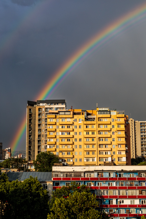 A double rainbow arcs over Soviet-era apartment buildings in Tbilisi, Georgia, with yellow and concrete façades lit beneath a dark, stormy sky.