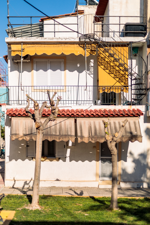 A residential building in Athens, Greece, features stacked balconies with yellow awnings, exterior staircases, and two pruned trees standing in front of the façade.