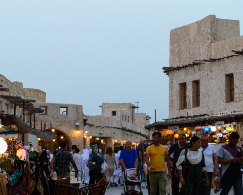 People walk through a busy market street in Doha, Qatar, lined with traditional buildings and shops illuminated by hanging lights.