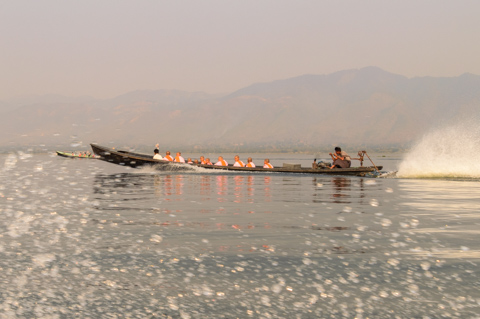 Burmese Buddhist nuns, known as thilashin, travel across Inle Lake in a long wooden boat, gliding over calm water with mountains faintly visible in the distance.