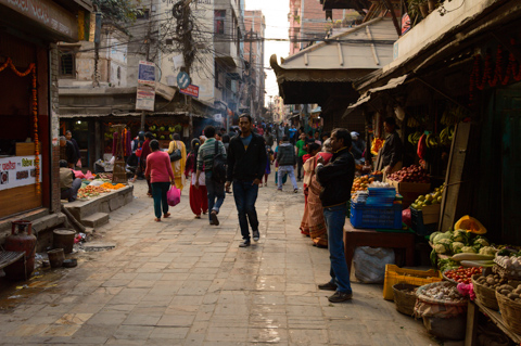 Pedestrians move through a narrow street lined with produce stalls and small shops in the heart of Kathmandu, Nepal, with baskets of fruits and vegetables displayed along the sidewalks.