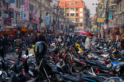 Rows of tightly packed motorbikes fill a crowded street in Kathmandu, Nepal, with pedestrians and shopfronts lining both sides.