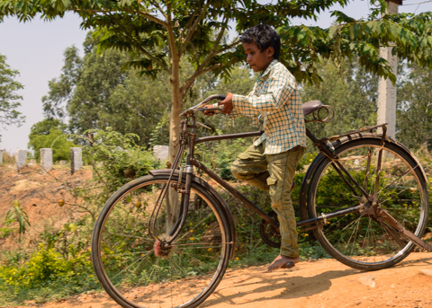 A young boy rides an adult-sized bicycle along a dirt path in Hampi, Karnataka, India, with one leg threaded through the frame beneath leafy trees.