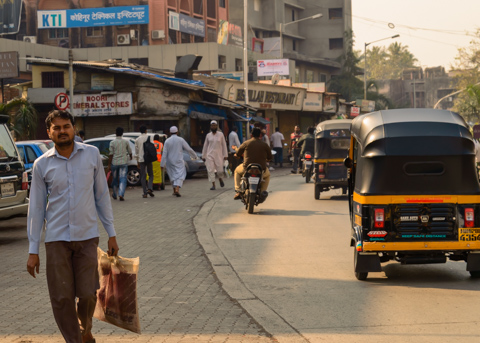Pedestrians, auto rickshaws, and motorcycles move along a busy street in Mumbai, India, as shops and traffic fill the late-afternoon scene.