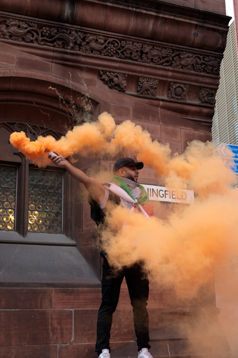 A man draped in a Palestinian flag releases orange smoke from a flare during a protest in Manchester, United Kingdom, standing against a stone building.