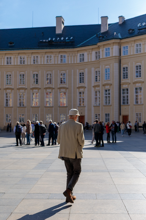 An elderly man wearing a beige hat and sports coat walks across the courtyard of Prague Castle, blending into the pale stone architecture in Prague, Czech Republic.