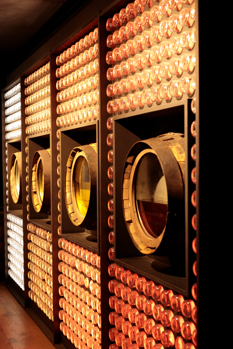 Four display barrels filled with whiskey of varying colors are set into a wall of backlit bottles at Teeling Distillery in Dublin, Ireland.