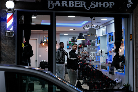 Street view of a Greek barbershop in Athens; two barbers look at the camera as the photo is taken.