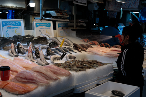 A fish vendor arranges fresh whole fish, fillets, and shrimp on crushed ice at an indoor seafood market in Athens, Greece, with handwritten price signs hanging above the display.