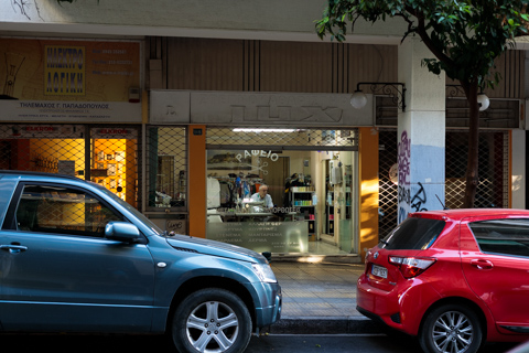 A tailor works inside a small street-level shop in Athens, Greece, seen through the open storefront between parked cars along a shaded city sidewalk.