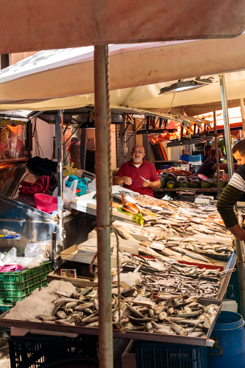 A fish vendor stands behind a stall piled with fresh fish at a busy laiki market in Athens, Greece, under canvas awnings.