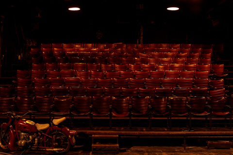 Rows of red vinyl seats sit empty in a dimly lit old theater in Athens, Greece, with a vintage motorcycle parked at the front.