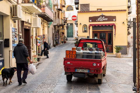 A small red utility truck loaded with metal canisters drives along a narrow cobblestone street in Cefalù, Sicily, as pedestrians walk past historic buildings in the old town.