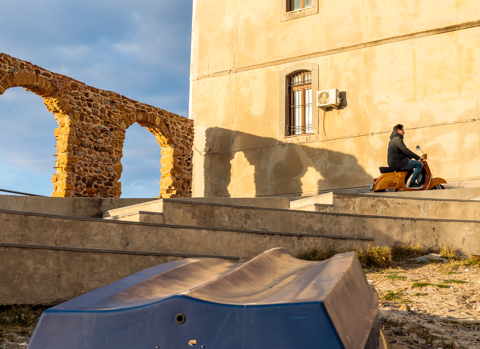 A man rides a Vespa scooter along a stepped stone walkway in Cefalù, Sicily, passing sunlit historic walls and the ruins of a stone arch under a clear sky.
