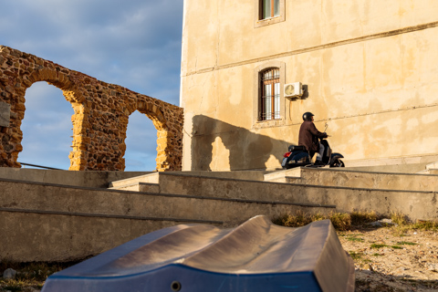 A man rides a Vespa scooter past sunlit stone arches and a weathered building in Cefalù, Sicily, Italy, with long shadows cast across the steps.