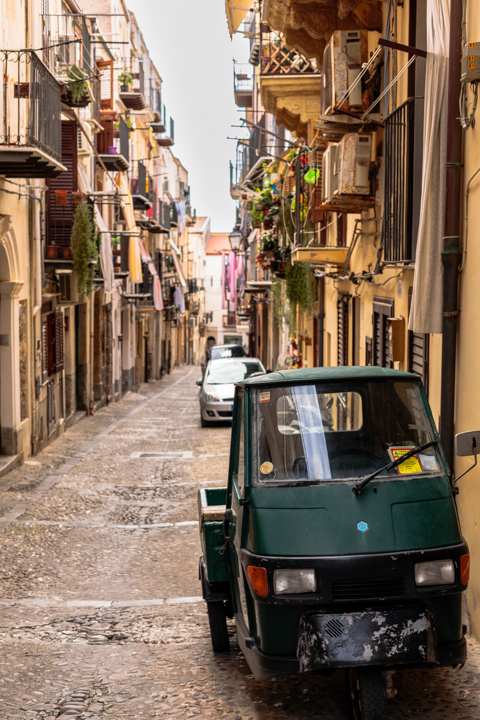 A small green three-wheeled utility vehicle is parked along a narrow cobblestone street in Cefalù, Sicily, lined with tall residential buildings and balconies filled with plants.