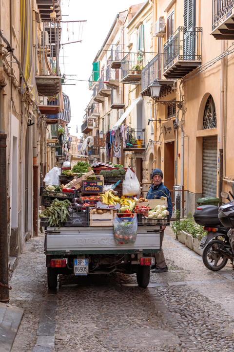 A produce vendor stands beside a small truck loaded with fruits and vegetables on a narrow cobblestone street in Cefalù, Sicily, Italy.