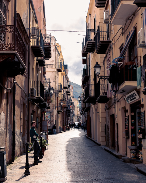 A narrow cobblestone street lined with weathered buildings and wrought-iron balconies stretches through the historic center of Cefalù, Sicily, Italy.